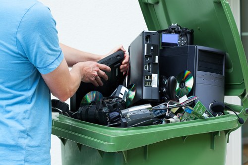 A person wearing a light blue T-shirt is shown in close-up, holding a portable electronic device while standing beside an open green waste disposal bin. Inside the bin, various discarded electronic items are visible, including a black desktop computer tower, a digital camera, and several CDs or DVDs scattered among other miscellaneous electronic waste. The scene appears to be outdoors or in a well-lit area, with the green bin positioned on a paved surface. The image emphasizes the collection and removal of electronic waste as part of rubbish clearance services, with a professional and neutral tone suitable for a garden clearance company operating in Soho and surrounding areas. The surrounding environment and background are not visible, focusing entirely on the contents of the bin and the person managing the waste.