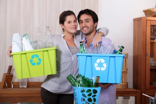 A young couple standing indoors in a room with light-colored wooden panels on the wall behind them, smiling at the camera while holding two plastic recycling bins filled with empty glass bottles and jars. The woman on the left, with dark hair tied back, is holding a bright green bin with a recycling symbol, containing clear and green glass bottles, some with caps. The man on the right, with dark curly hair and a beard, is holding a blue bin also marked with a recycling symbol, filled with similar glass bottles and jars, some with caps and labels. In front of the couple, a smaller blue plastic container holds additional green glass bottles. The setting appears to be a home environment focused on recycling and rubbish separation, possibly in Soho, with a wooden cabinet partially visible to the right. The image conveys a sense of engagement with waste sorting and recycling activities at home, aligned with rubbish removal services for domestic or commercial premises in the area.