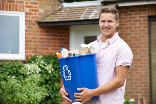 A young man with short, light brown hair and a friendly smile is standing outdoors in front of a brick house with a white window and a small front garden featuring green shrubs and white flowers. He is wearing a pale pink short-sleeved shirt and is holding a blue recycling bin filled with various types of rubbish, including paper, cardboard, and plastic items. The scene is well-lit with natural daylight, and the background suggests a residential area typical of Soho. This image is related to rubbish removal and garden clearance services offered by Garden Clearance Soho, serving local areas and postcode districts in the region. The man appears to be preparing to move the waste for disposal or collection, emphasizing a professional and practical approach to rubbish clearance.