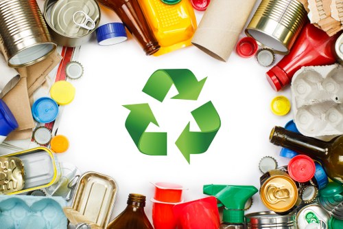 An assortment of discarded recyclables, including aluminum cans, glass bottles, plastic containers, and cardboard tubes, arranged on a flat surface surrounding a large green recycling symbol at the centre. The cans are mostly crushed or whole with metallic finishes in silver, gold, and painted colours, while the plastic bottles are in various shades such as red, blue, and yellow, with some caps attached. Cardboard tubes and pieces of paper are also visible among the waste items. The background is a neutral white, highlighting the vibrant colours of the packaging materials. This image represents waste segregation and recycling efforts commonly seen in garden clearance and rubbish removal services, such as those provided by Garden Clearance Soho in the Soho area, as part of responsible waste disposal practices.