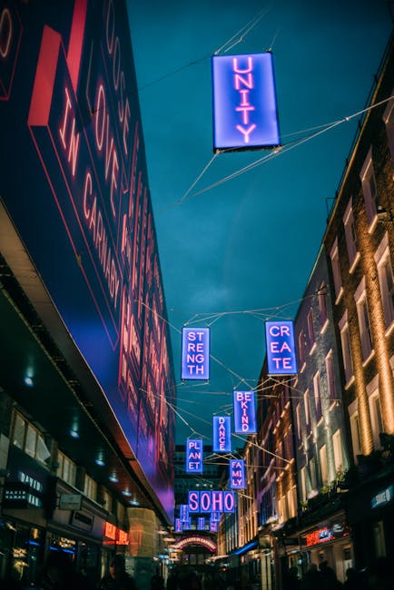 A bustling city street scene in Soho with pedestrians walking along the pavement on both sides of the narrow road. The street is lined with multi-story buildings featuring a mix of architectural style