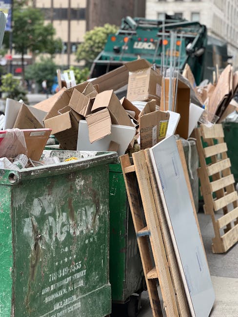 A large, green metal dumpster with a weathered surface, showing rust and faded white paint, is positioned outdoors on a paved street or sidewalk. The open top reveals a variety of waste materials incl