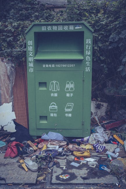 A pile of black rubbish bags, some torn open, is stacked in front of a red metal door on an exterior wall with a rough, light-colored textured surface. The bags contain various waste materials and ref